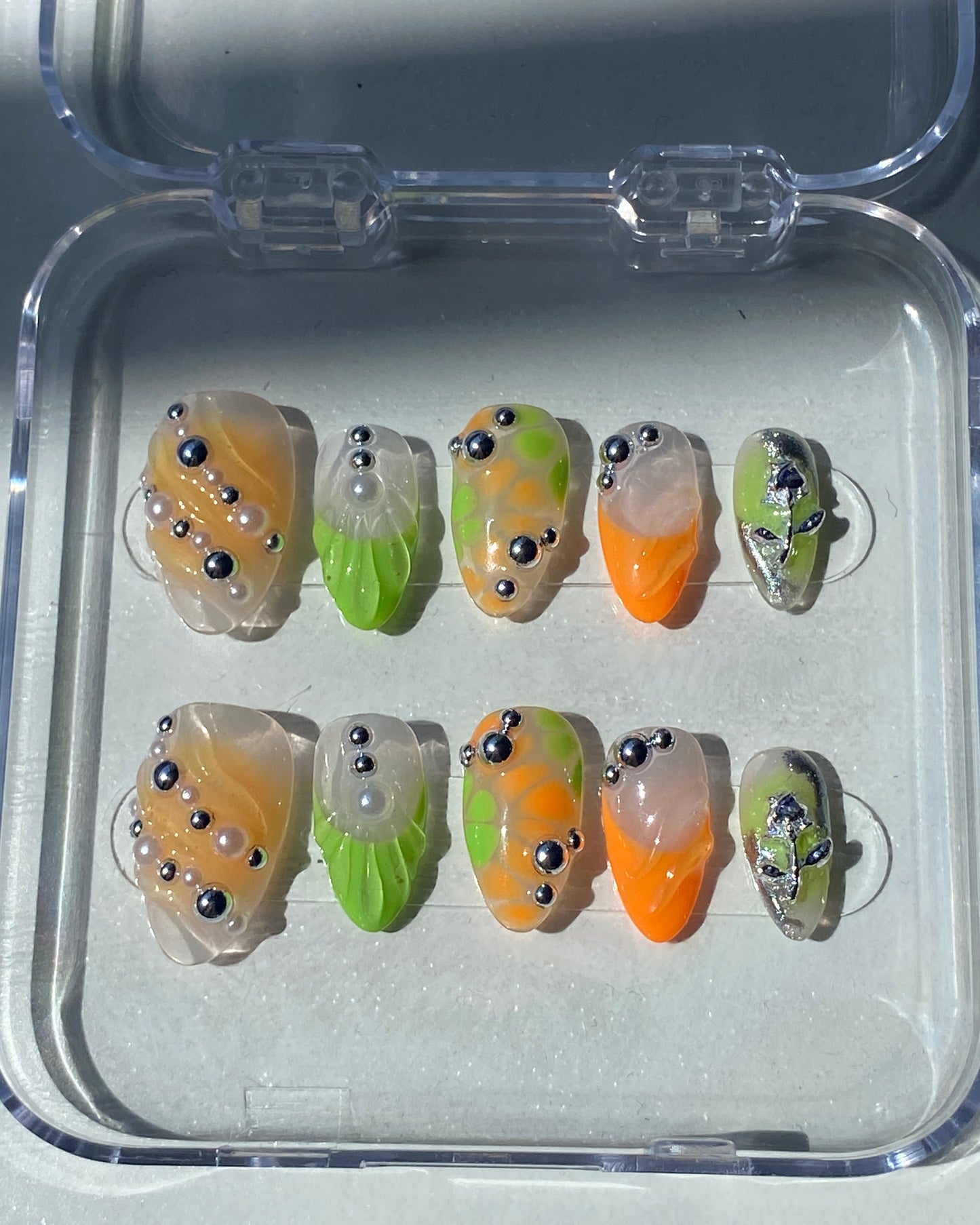 A set of ten abstract press-on nails is displayed in a clear case under direct sunlight. The designs feature translucent peachy beige, neon green, and orange sections, accented with silver and pearl-like studs and 3D sculptural elements, including a silver alien face on one nail.