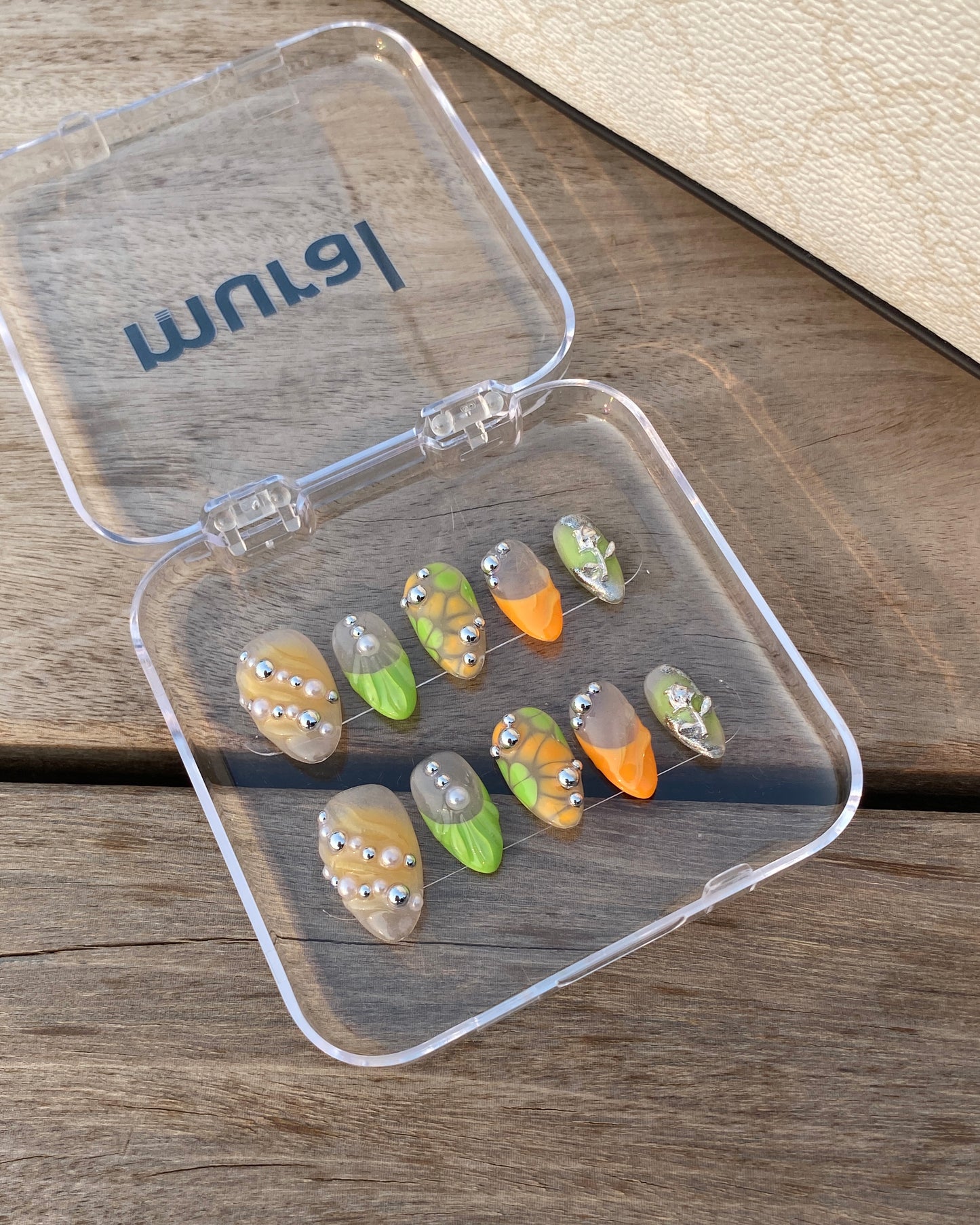The Sweet Tang nails are arranged in a clear mural-labeled case on a wood surface with warm lighting. The varied designs feature bright citrus-inspired colors, chrome embellishments, and one standout silver alien nail.