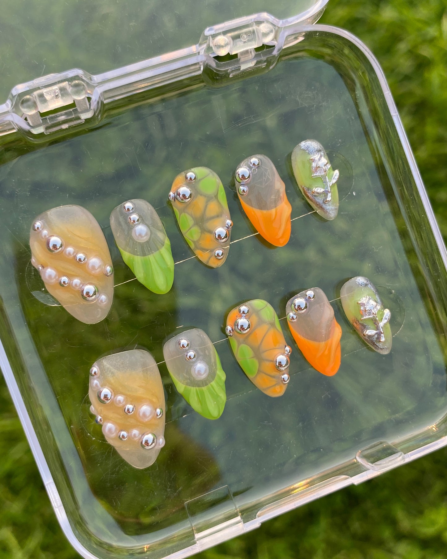 The Sweet Tang nail set is shown from above in a clear box placed on green grass. Each nail features vibrant, abstract designs in orange, green, and nude tones, embellished with silver and pearl beads, including textured sculptural patterns and a metallic alien detail.