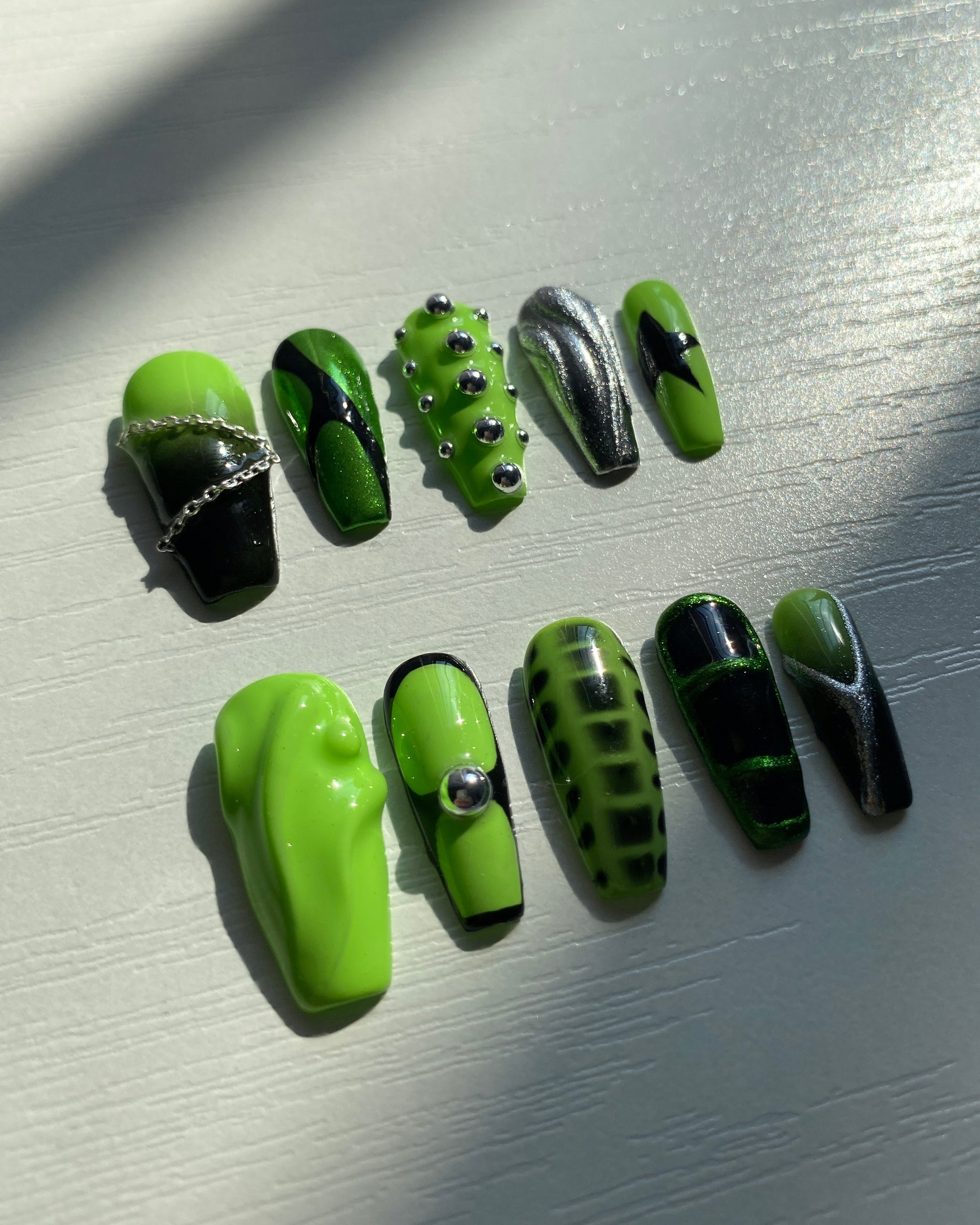 Y2K-inspired green and black press-on nails catching natural light, showcasing high-shine finishes, chrome texture, and bold 3D silver studs.