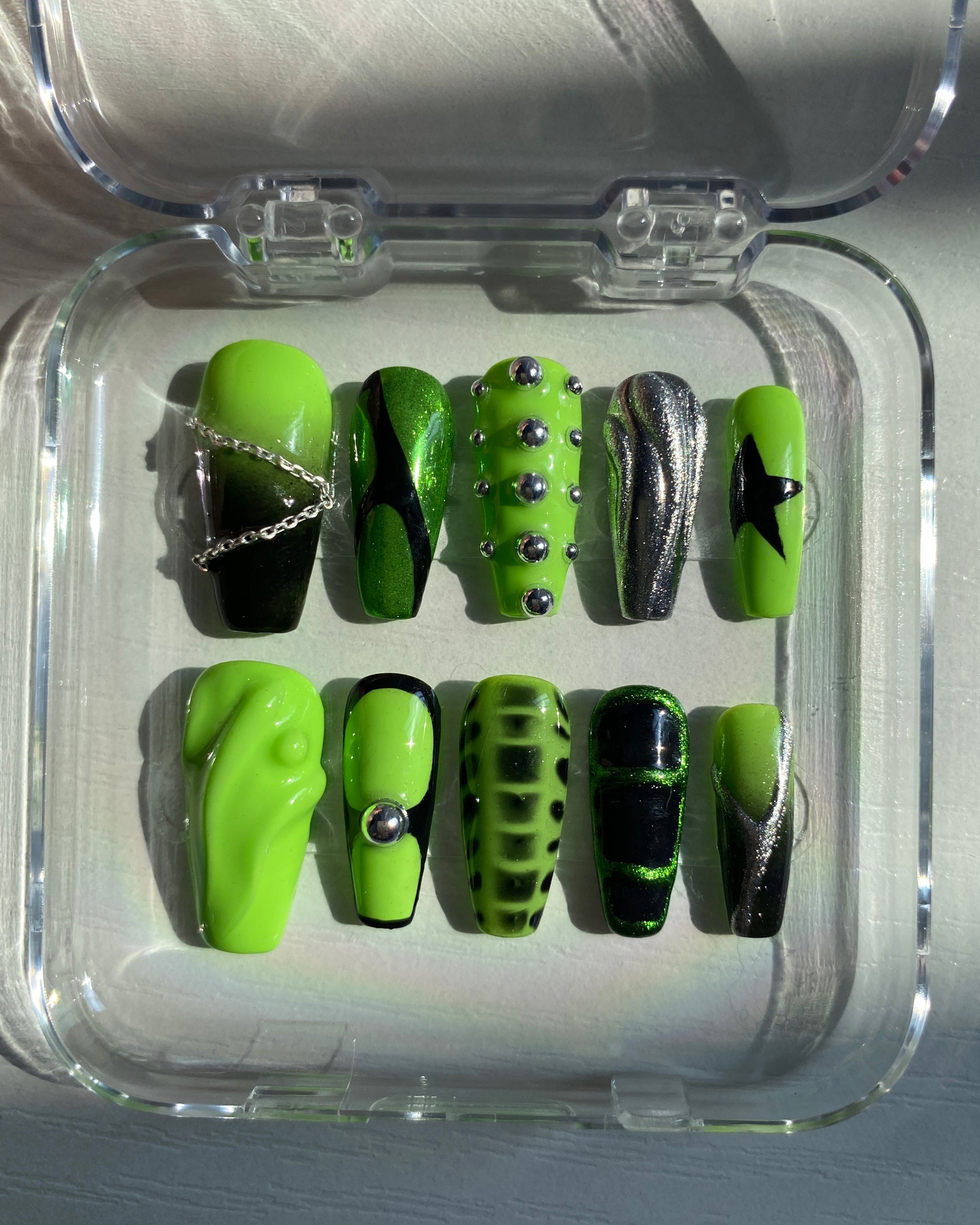 Neon green and black press-on nails neatly displayed in a clear case, featuring Y2K designs with metallic elements, chains, and bold abstract patterns.