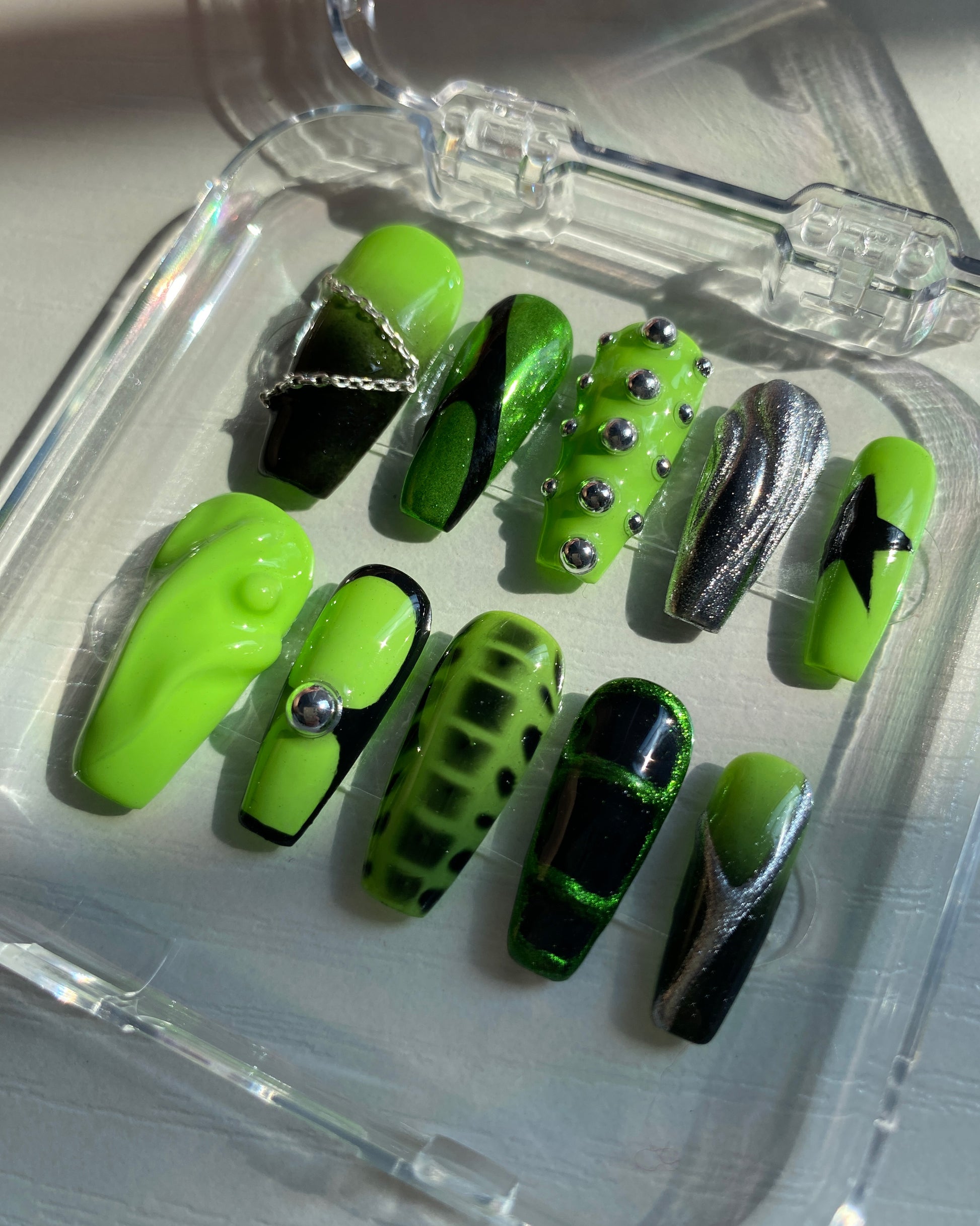 Angled view of a press-on nail set inside a clear box, highlighting Y2K nail art with 3D silver studs, swirl patterns, and eye-catching neon green and chrome color palette.