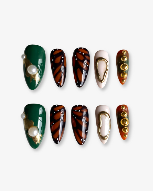 Overhead view of the full nail set laid out on a white crisp background, capturing the intricate mix of textures and finishes: glossy, metallic, and pearlescent elements with monarch butterfly motifs, minimalist gold designs, and earthy tones.