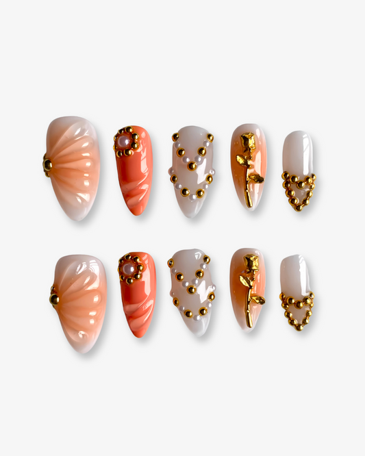 The Poetic press-on nail set is laid out against a white background, showcasing symmetrical designs in warm coral and nude tones. Each nail includes ornate golden details, from a sculpted rose to pearl-centered clusters and gold-beaded hearts.
