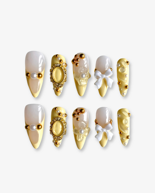 A set of ten long almond-shaped press-on nails with intricate 3D embellishments, featuring a yellow base and pinkish nude top. Designs include pearl clusters, gold studs, ornate gold frames, sculpted white bows, and floral elements.