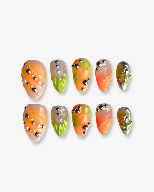 An image of the Sweet Tang press-on nail set on a white background shows ten uniquely designed nails with swirling orange and nude tones, bright green leafy textures, polka-dot patterns, and shiny silver accents—each with a bold and playful 3D effect.